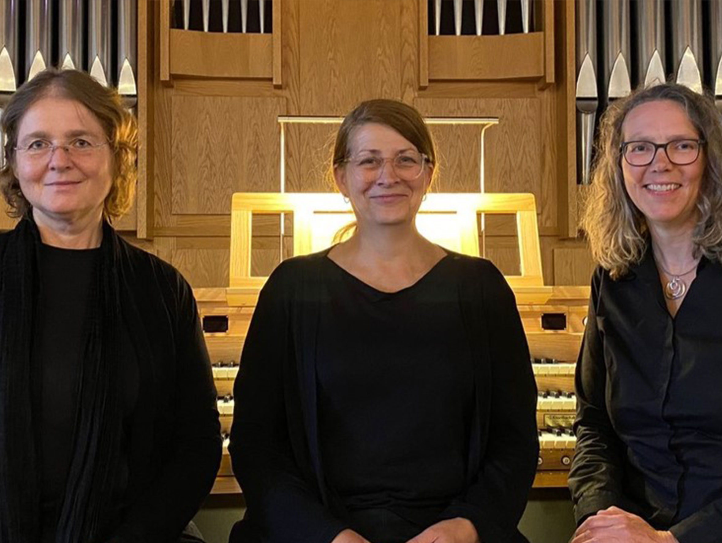 Drei Frauen in Schwarz, lächelnd, vor einer Orgel.