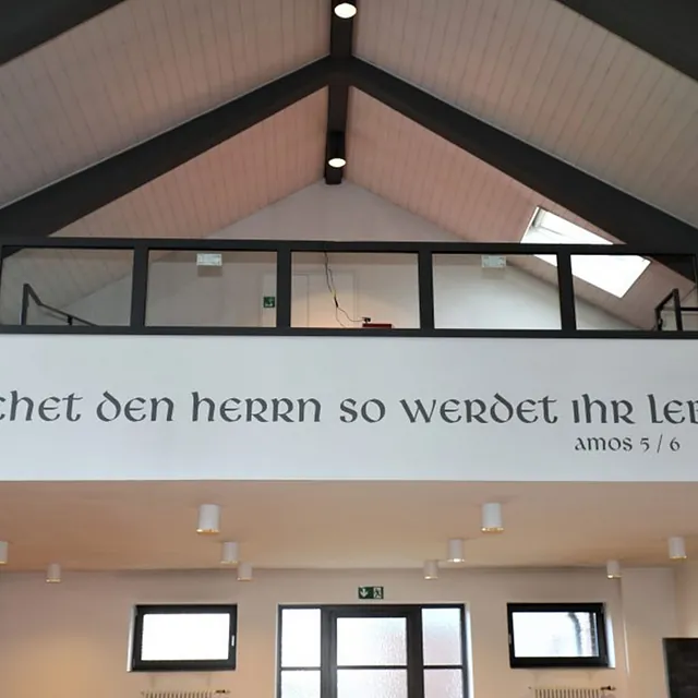 Kircheninnenraum: Deutscher Schriftzug "Amos 5/6" an Wand, hohe Decken.