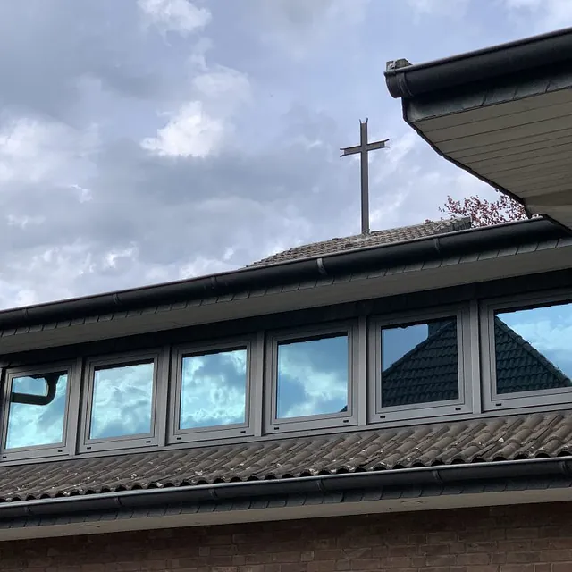 Kirchendachlinie mit himmelreflektierenden Fenstern und einem Kreuz.