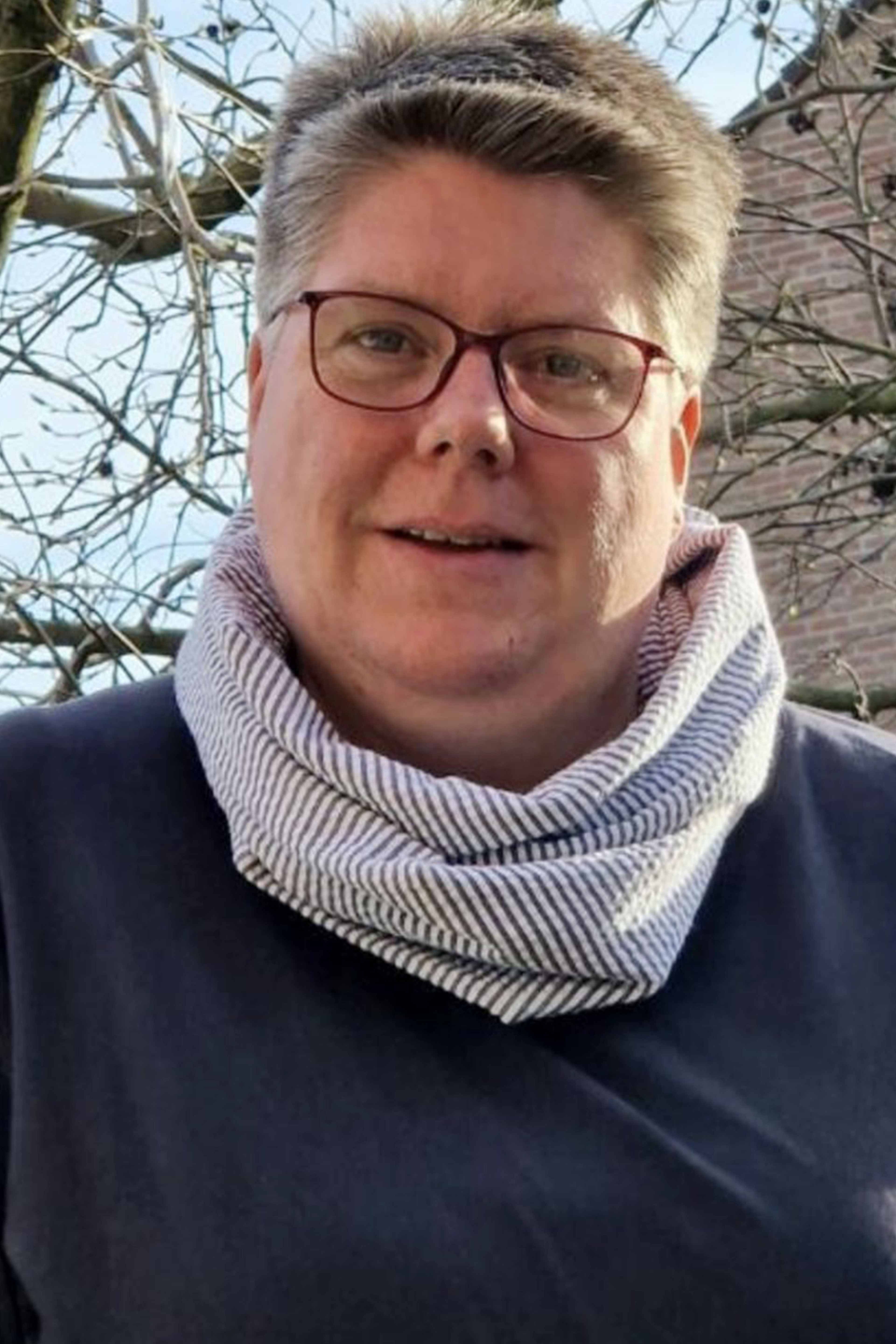 Person mit kurzen Haaren, Brille, Schal, lächelt draußen. Lässig.