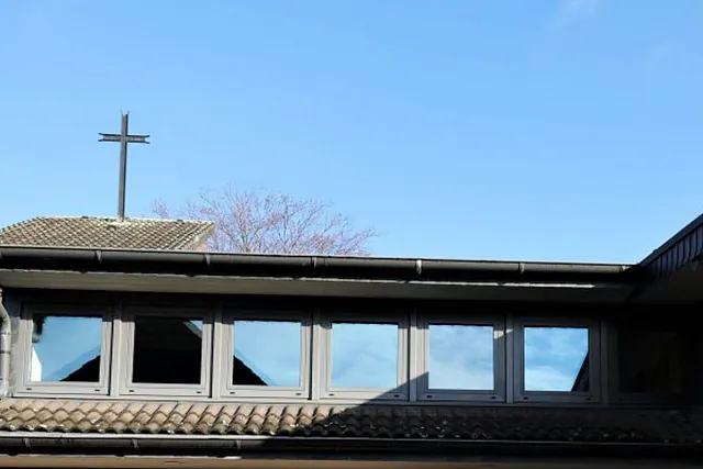 Kirchendach mit Kreuz und Fenstern vor blauem Himmel.