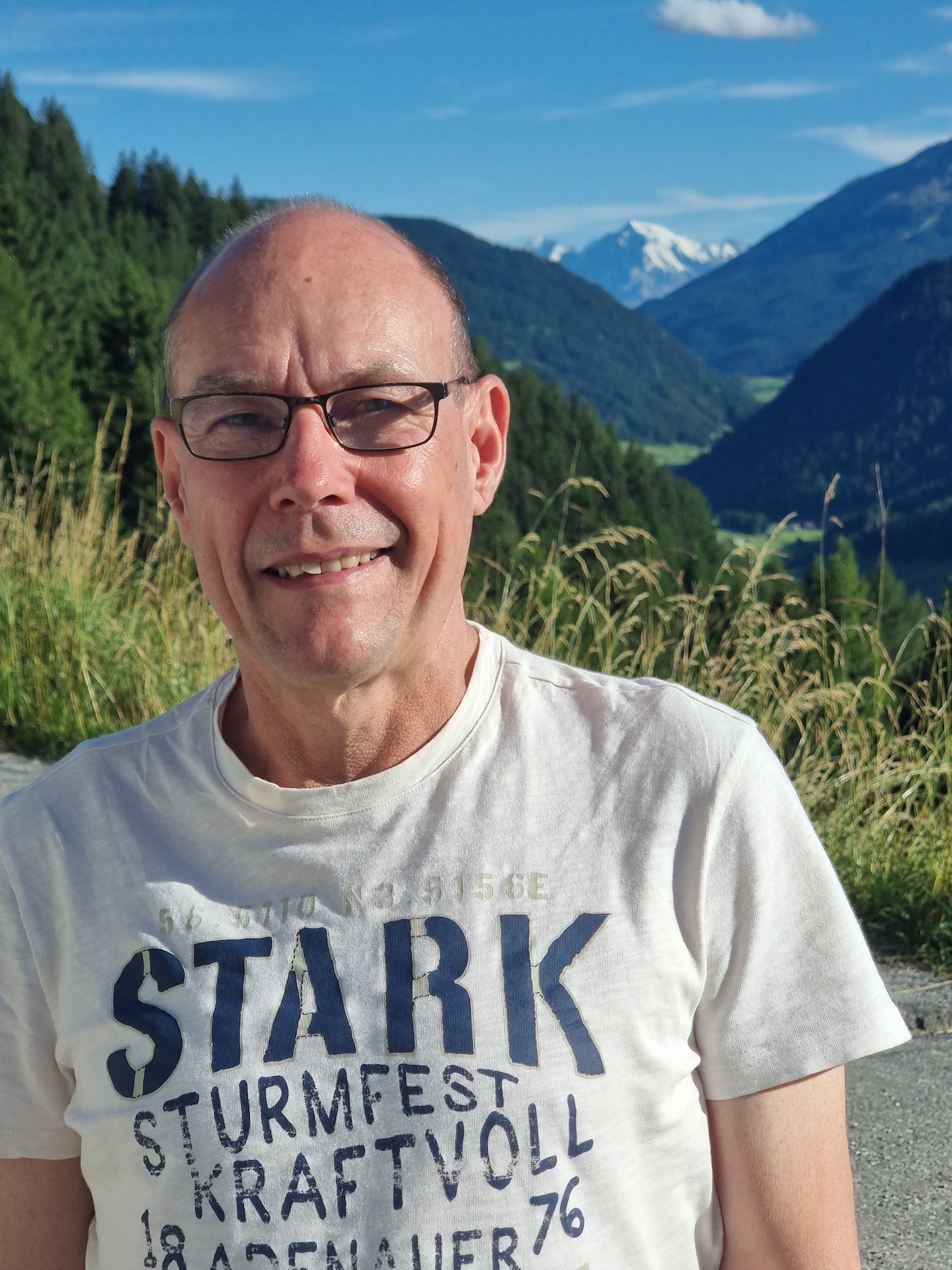 Person mit kurzen Haaren, Brille, Schal, lächelt draußen. Lässig.
