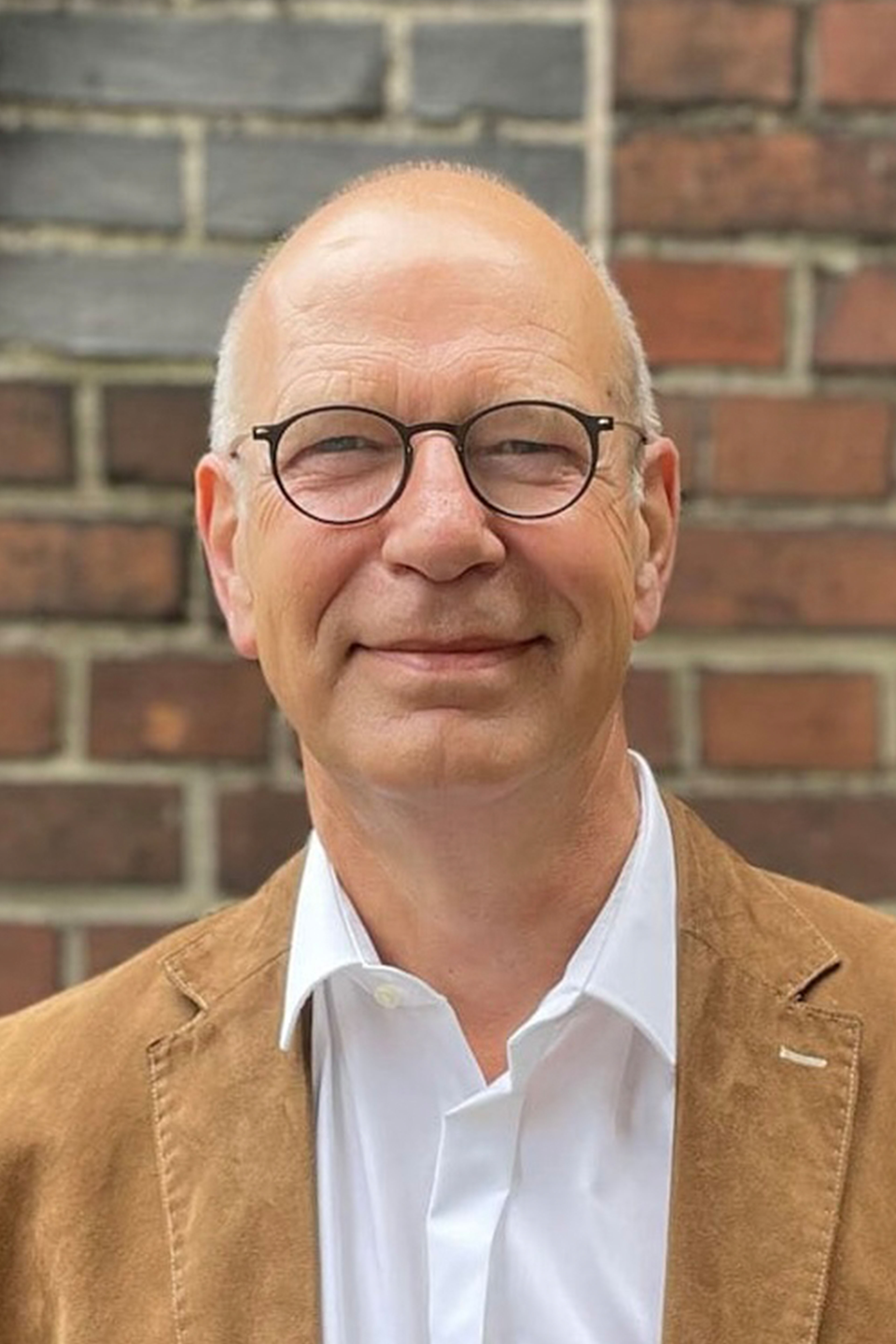 Lächelnder Glatzkopf mit Brille und brauner Jacke, Ziegelhintergrund.