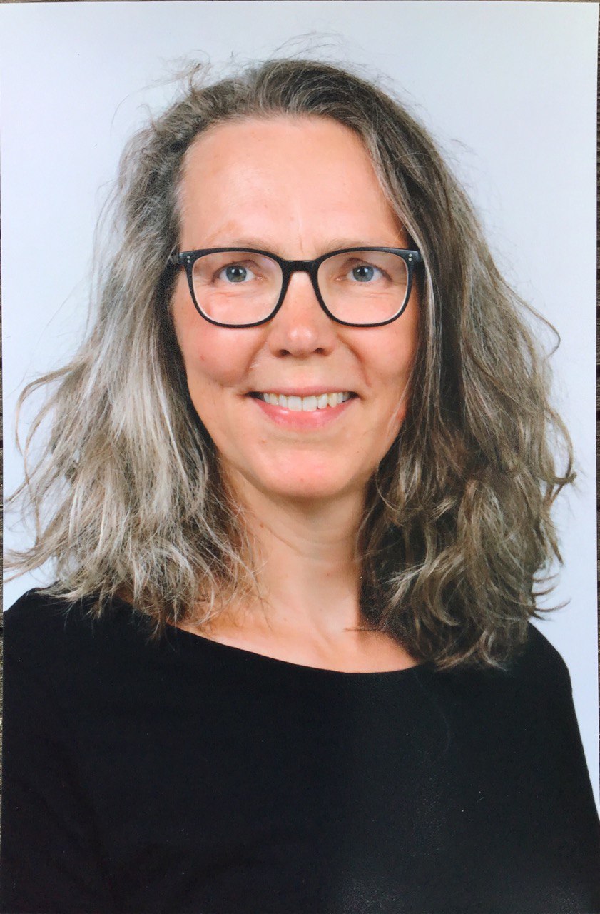 Eine lächelnde Frau mit grauem Haar und Brille, die ein schwarzes Oberteil trägt.